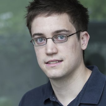 © MPIfG / Dünkelmann A young man with short dark hair and glasses, wearing a navy blue polo shirt, looking at the camera.