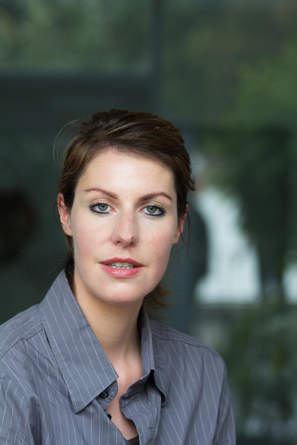 A woman with short brown hair and green eyes, wearing a gray button-up shirt, looking at the camera.