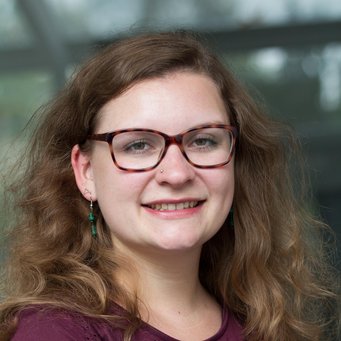 © MPIfG /Dünkelmann A smiling person with curly hair, wearing glasses and earrings, posing for a portrait.