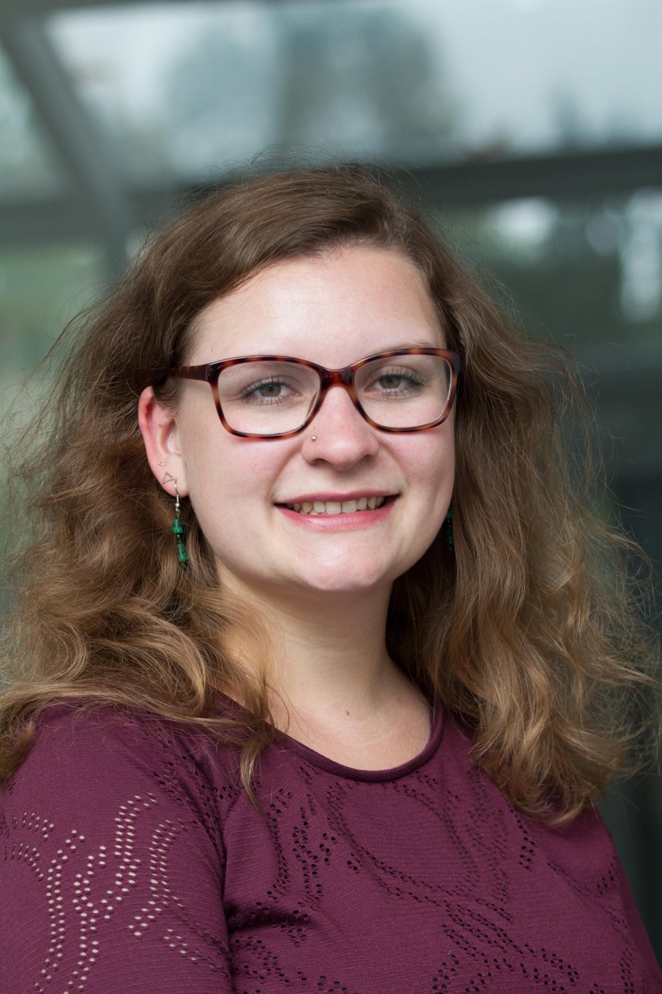 A smiling person with curly hair, wearing glasses and earrings, posing for a portrait.