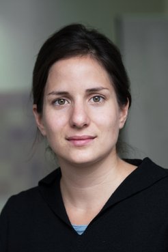 A woman with light brown skin and dark hair is looking directly at the camera, wearing a black top.