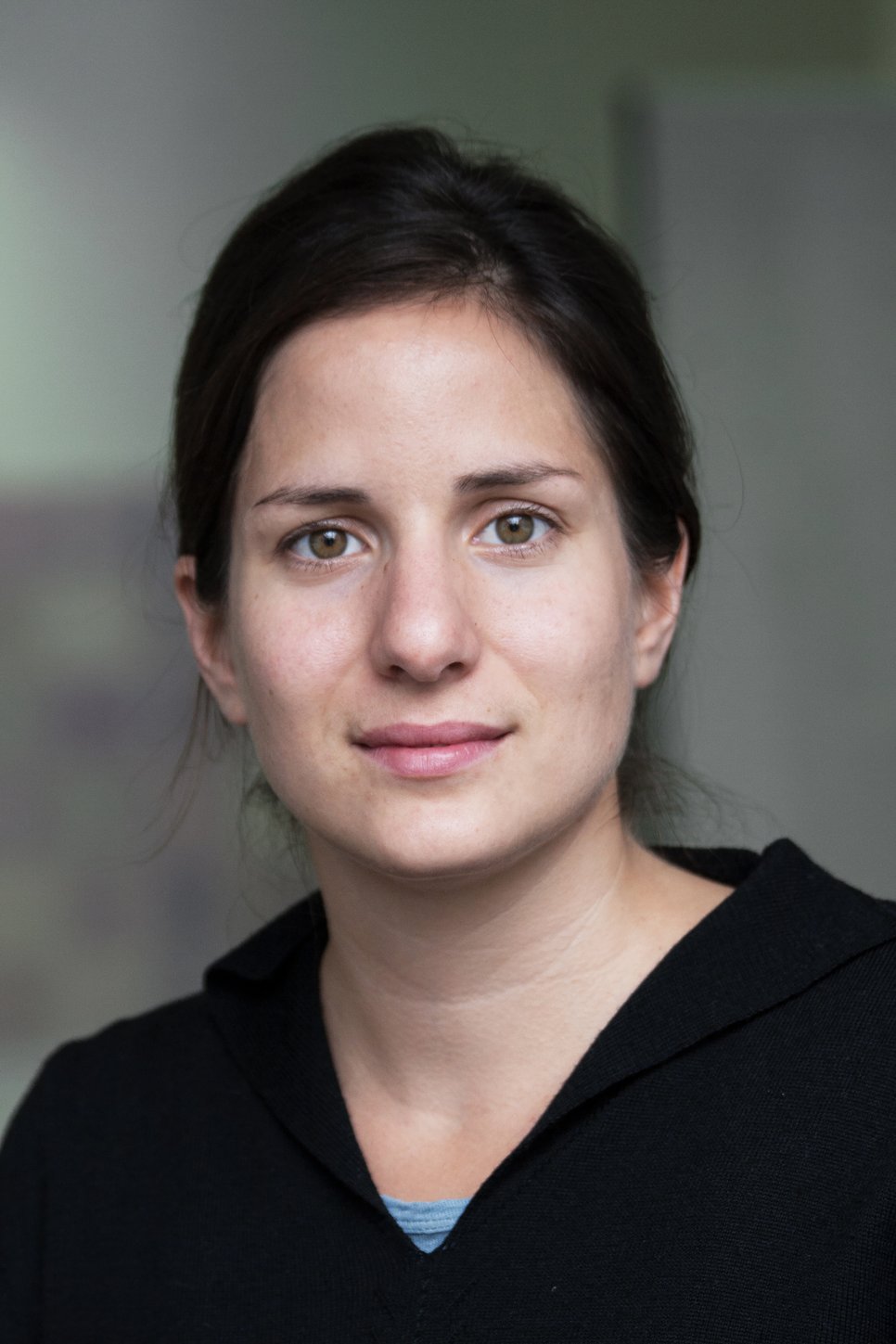 A woman with light brown skin and dark hair is looking directly at the camera, wearing a black top.