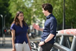 A woman in a blue shirt and a man in a dark shirt are smiling and engaging in conversation on a sunny day near a railing.
