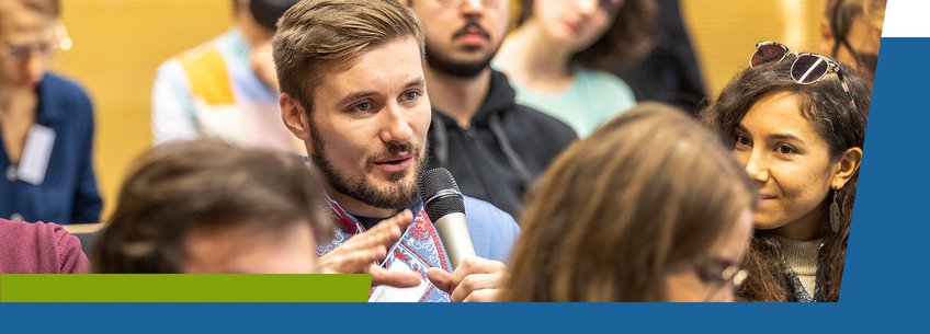 A man speaking into a microphone at a conference A man speaking into a microphone at a conference or event, surrounded by an audience.