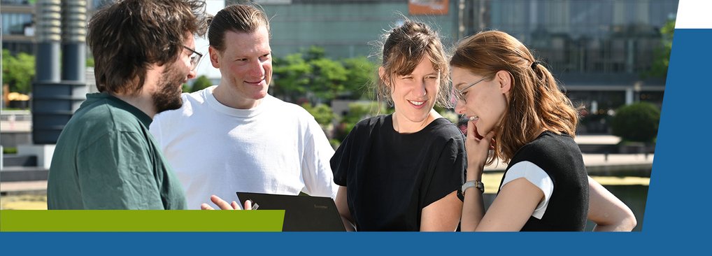 A group of four people engaged in conversation outdoors, standing together and looking at a laptop.