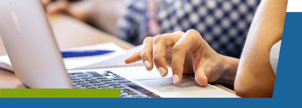 A person’s hand is using a laptop keyboard, with a notebook and pen visible in the background.
