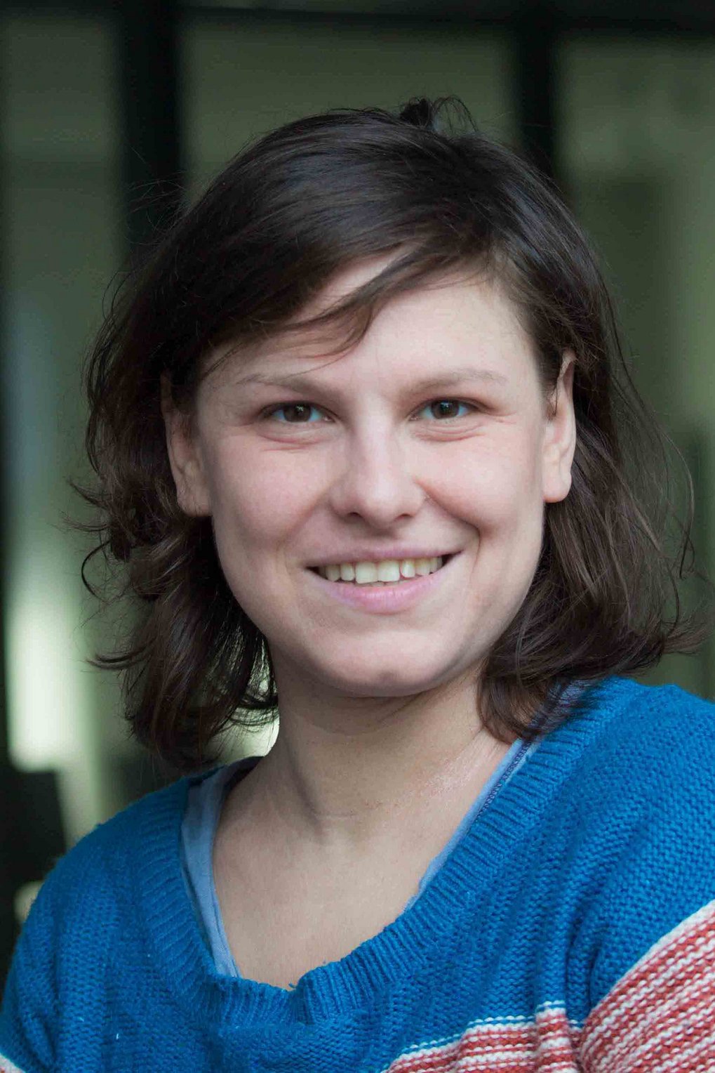 A close-up portrait of a person with medium-length brown hair, smiling and wearing a blue sweater.
