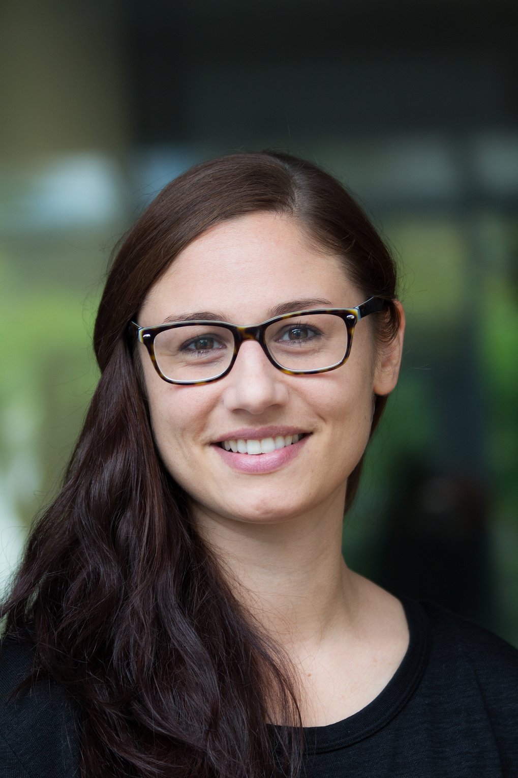 A smiling woman with long dark hair and glasses, set against a blurred background.