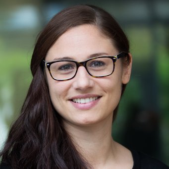 A smiling woman with long dark hair and glasses, set against a blurred background.