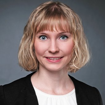 A woman with short, wavy blonde hair and blue eyes is smiling at the camera, wearing a black jacket over a white shirt. The background is gray.