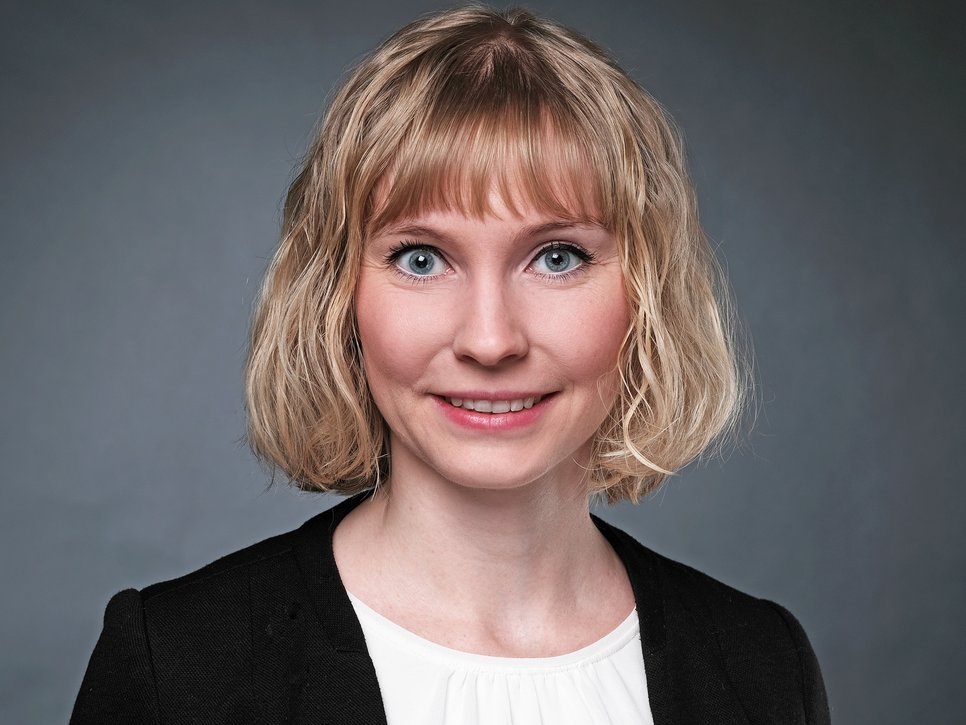 A woman with short, wavy blonde hair and blue eyes is smiling at the camera, wearing a black jacket over a white shirt. The background is gray.