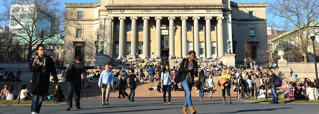 A busy scene outside the Library of Columbia University, featuring people sitting on the steps and walking in front, with the building's columns visible in the background.