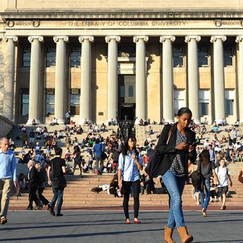 A busy scene outside the Library of Columbia University, featuring people sitting on the steps and walking in front, with the building's columns visible in the background.