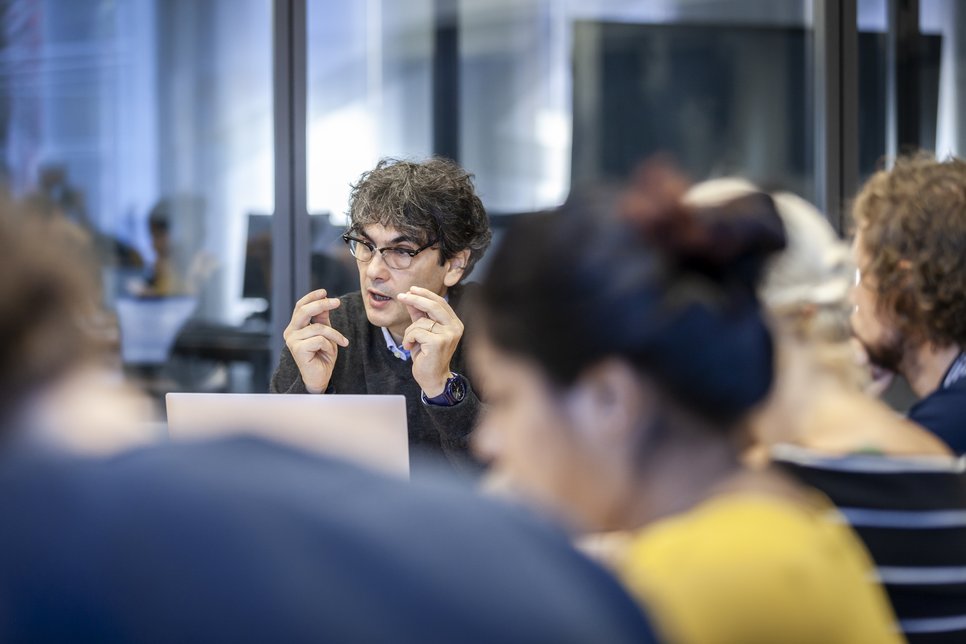 Group of people engaged in a meeting, focus on speaker gesturing.