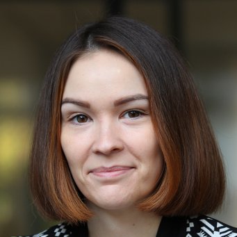 A close-up portrait of a woman with shoulder-length brown hair, smiling, wearing a patterned black and white jacket.