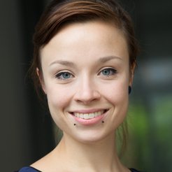 A close-up portrait of a smiling woman with brown hair and blue eyes, wearing a blue top and two facial piercings.