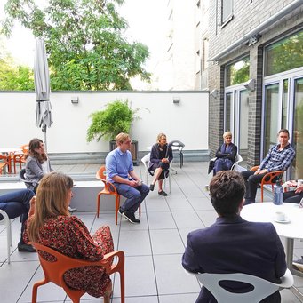 A group of people sitting in a circle on a patio, engaged in conversation, surrounded by greenery and modern furniture.