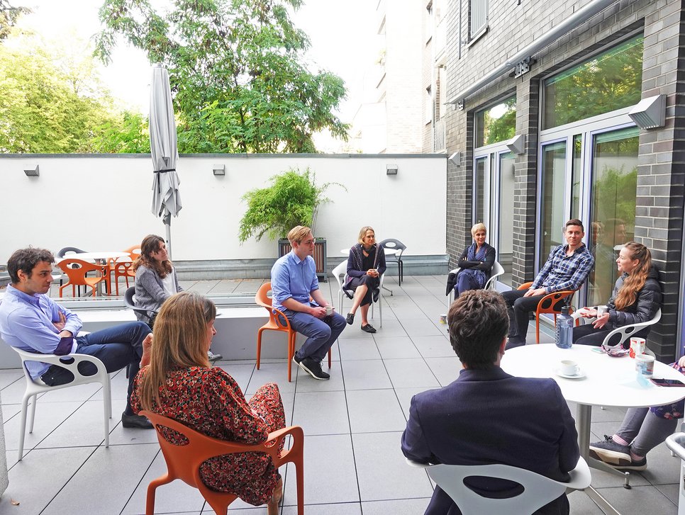 A group of people sitting in a circle on a patio, engaged in conversation, surrounded by greenery and modern furniture.